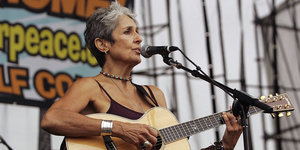 Eine ältere Frau mit kurzen, dunkelgrauen Haaren spielt Gitarre und singt in eine Mikrofon