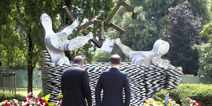 Zwei Männer von hinten vor einem Denkmal und vielen Blumenkränzen