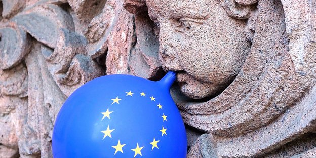Luftballon in EU-Blau mit Sternen wird vom Mund einer Steinputte aufgeblasen