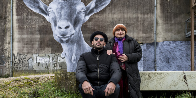 Die Regisseurin und der Künstler sitzen auf einer Böschung im Gras, dahinter ist das Wandbild einer Ziege.