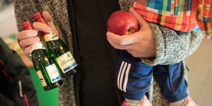 Eine Person hält zwei Flaschen Sekt und einen Apfel in der Hand. Im Arm hat sie ein Kind
