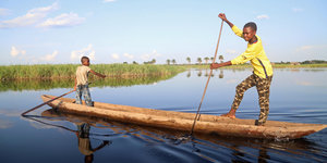 Zwei Kinder, die in einem Boot stehen und sich mit Stöcken vom Grund des Gewässers abstoßen. Die Landschaft dahinter ist flach