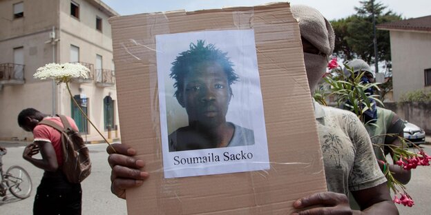 Maskierter Mann hält Karton mit Foto von Sacko Soumali in die Kamera