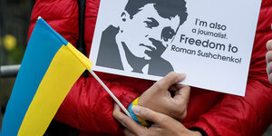 Eine Person hält eine ukrainische Flagge und ein Schild in der Hand mit der Aufschrift „I'm also a journalist. Freedom to Roman Sushchenko!““