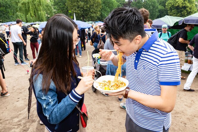 Zwei Menschen essen im Stehen mit Stäbchen von einem Plastik-Teller