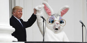 Trump hält im Weißen Haus die Hand eines Menschen im weißen Osterhasenkostüm hoch.