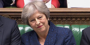 Theresa May, eine Frau mit kurzen grauen Haaren lächelt. Sie sitzt auf einer grün gepolsterten Bank. Eine Frau mit grauen kurzen Haaren spricht in ein Mikrofon, im Hintergrund gucken viele Menschen gespannt zu ihr