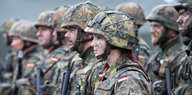 Mehrere Soldaten und Soldatinnen stehen in Reihen. Sie haben Helme und Uniform an und sind mit Farbe im Gesicht angemalt