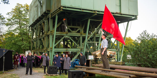 Ein Mann steht vor einer Industrieanlage und hält eine rote Fahne in die Luft