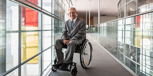 Wolfgang Schäuble, ein deutscher Politiker im Rollstuhl, wartet in einem Gang und lächelt.