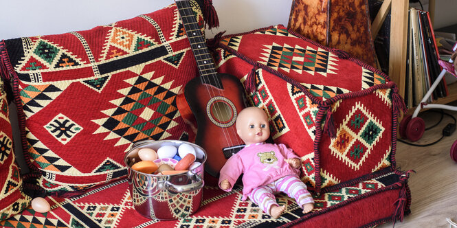 Ein Sofa mit ornamentvollem Bezug. Darauf sind eine Puppe, eine Gitarre und ein Topf, in dem Eier und Spielsachen sind