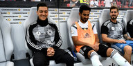 Die Fußballspieler Mesut Özil, İlkay Gündoğan und Kevin Trapp (von links nach rechts) sitzen in Sportkleidung nebeneinander auf der Bank