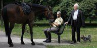 Ein Polizist sitzt auf einer Parkbank und hält ein Pferd am Halfter fest