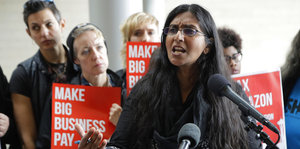 Seattles Stadträtin Kshama Sawant spricht. Im Hintergrund Menschen mit Schildern, auf denen steht: „Make Big Business Pay"