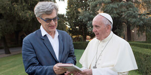 Papst Franziskus und Wim Wenders stehen in einem Garten und halten ein Buch