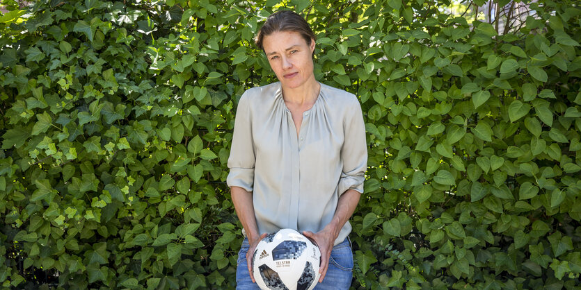 Katja Kraus steht vor einer Hecke und hält einen Fußball in den Händen