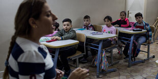 Kinder sitzen auf Schulbänken, vorne die Lehrerin