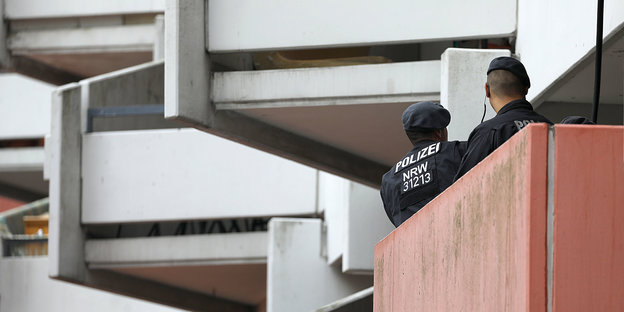 Zwei Polizisten sind von hinten auf einem hornhautfarbenen Balkon zu sehen, über ihnen türmen sich weiße Beton-Balkone