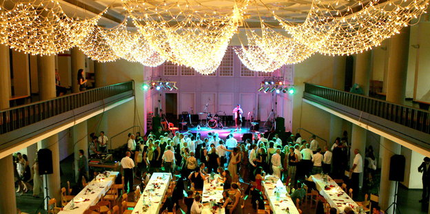 In einem großen und edel geschmücktem Saal mit Esstischen tanzen Menschen vor einer Bühne