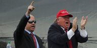 Zwei Männer auf einer Tribüne: Einer reckt die rechte Faust in Siegrpose, daneben klatscht ein mann, der wie der US-Präsident Tump aussieht