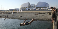 Junge Männer springen am Strand von Sotschi ins Wasser. Im Hintergrund befindet sich das WM-Stadion.