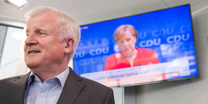 Horst Seehofer blickt nach links, hinter blickt Angela Merkel von einem Fernsehbildschirm herunter
