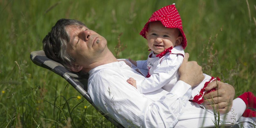 Ein Mann liegt auf einer Wiese in einem Liegestuhl, auf ihm ein Baby mit roter Mütze