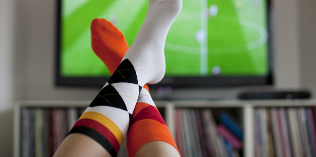 Ein Fernseher. Davor zu sehen: zwei Füße mit Socken in schwar-rot-gold