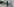 Eine Frau mit langen Kleid und einer Aldi-Plastiktüte in der Hand geht an einer Häuserfront vorbei; im Hintergrund sind unscharf drei junge Männer, vor einem Eingang stehend, zu sehen.