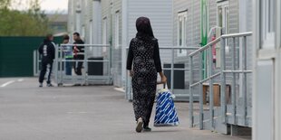 Eine Frau mit langen Kleid und einer Aldi-Plastiktüte in der Hand geht an einer Häuserfront vorbei; im Hintergrund sind unscharf drei junge Männer, vor einem Eingang stehend, zu sehen.