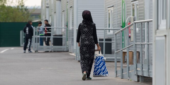 Eine Frau mit langen Kleid und einer Aldi-Plastiktüte in der Hand geht an einer Häuserfront vorbei; im Hintergrund sind unscharf drei junge Männer, vor einem Eingang stehend, zu sehen.