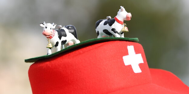 zwei kleine Plastikkühe sind auf einem roten Hut mit weißem Kreuz festgeschraubt