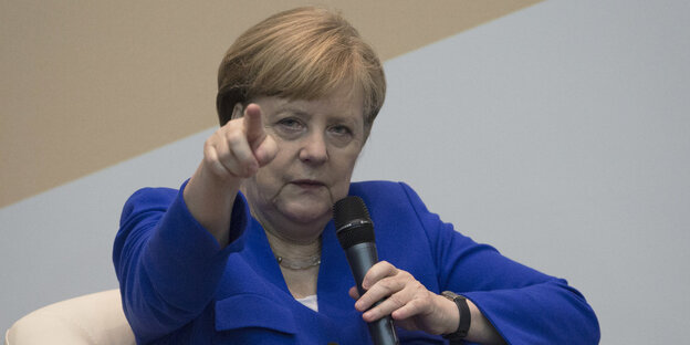 Angela Merkel sitzt zurückgelehnt in einem Sessel und zeigt mit dem rechten Zeigefinger nach vorne