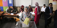 Eine Frau mit Kopftuch wirft ihren Wahlzettel in eine Wahlurne, hinter ihr warten weitere Menschen auf ihre Stimmabgabe
