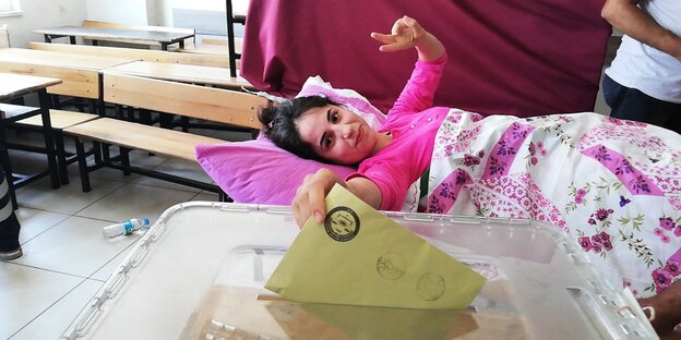 Eine junge Frau auf einer Krankenliege zeigt das Peace-Zeichen, während sie ihren Stimmzettel in eine Wahlurne wirft Eine junge Frau auf einer Krankenliege zeigt das Peace-Zeichen, während sie ihren Stimmzettel in eine Wahlurne wirft