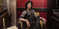 Patsy l’Amour laLove sitzt in einem Sessel und trägt ein schwarzes, trägerloses Kleid, eine Perlenkette und eine schwarze Perücke.