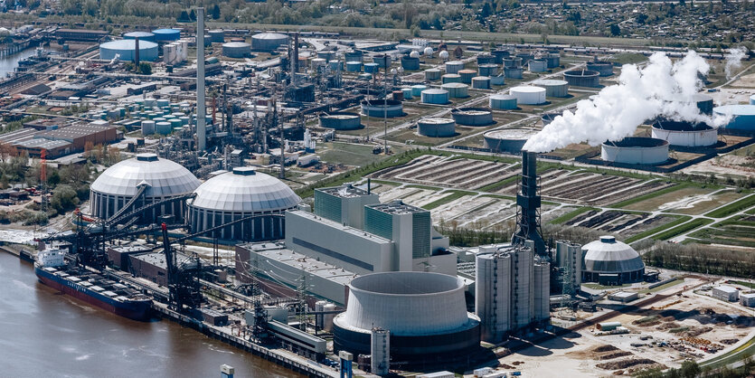 An einem Fluss steht ein großes Kraftwerk mit zwei runden Gebäuden und einem Schornstein, aus dem weißer Rauch kommt