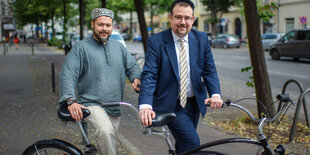 Vor einem großen Tandem-Fahrrad steht ein Mann mit einer muslimischen Kopfbedeckung, daneben ein Mann mit einer jüdischen Kopfbedeckung