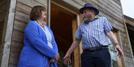 Frau im blauen Kleid und Mann mit Hut vor Holzhaus