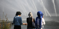 Drei Mädchen mit Kopftüchern stehen vor Wasserfontänen