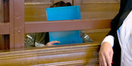 Ein Mann auf einer Bank im Gerichtssaal, wie er sich eine blaue Mappe vor das Gesicht hält; neben ihm steht eine Frau mit weißer Bluse, schwarzer Jacke und rot lackierten Nägeln