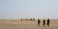 Blick über eine Wüstenlandschaft - im Vordergrund laufen Menschen, im Hintergrund das Skelett eines unfertigen Gebäudes