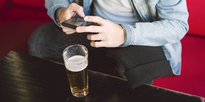 Ein Glas Bier auf einem Tisch, daneben Hände an einem Smartphone