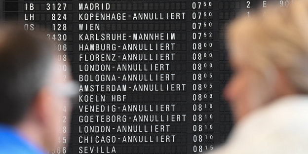Zwei Reisende blicken auf eine Anzeigetafel am Flughafen, auf der zahlreiche annullierte Flüge stehen