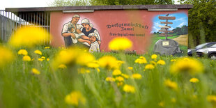 Auf einer Wiese blüht gelber Löwenzahn, dahinter steht auf einer braun-roten Scheunenwand in schwarzen, runenartigen Buchstaben: Dorfgemeinschaft Jamel - frei sozial national