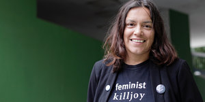 Sarah Ahmed steht vor einer grünen Wand und lächelt. Auf ihrem blauen T-Shirt steht in weißer Schrift „feminist killjoy“.