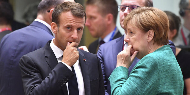 Merkel und Marcron stehen zusammen, beide haben die rechte Hand ans Kinn gelegt