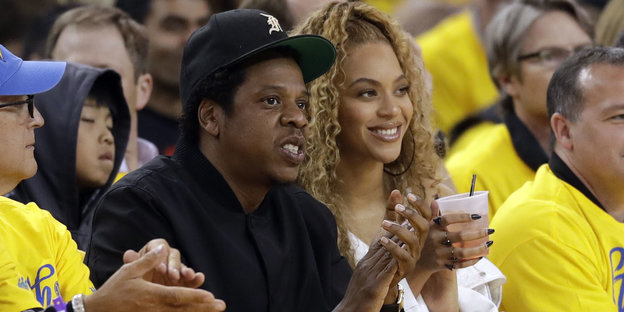 Jay-Z und Beyoncé sitzen nebeneinander in einem Basketball-Stadion und trinken Bier