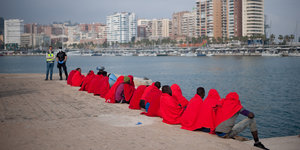 Menschen sitzen in rote Decken gehüllt an einem Hafenbecken