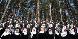 Ein Chor steht vor hohen Kiefern, alle sind schwarz gekleidet und haben das Haupt mit weißen Tüchern bedeckt